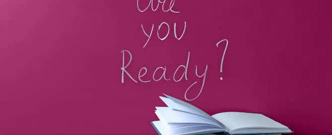 A desk with some work related items on it, with a sign on the wall saying "Are you ready?"