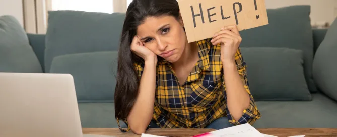 A woman is working at her desk, but looks disgruntled and holds up a sign that reads "Help"