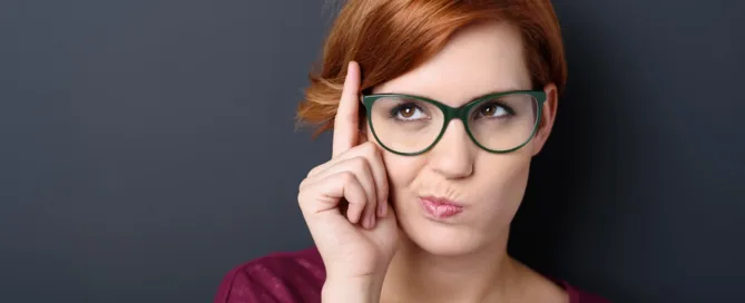 A lady wearing glasses sits thinking, whilst looking up to her right and holding her finger to her head indicating she is deep in thought